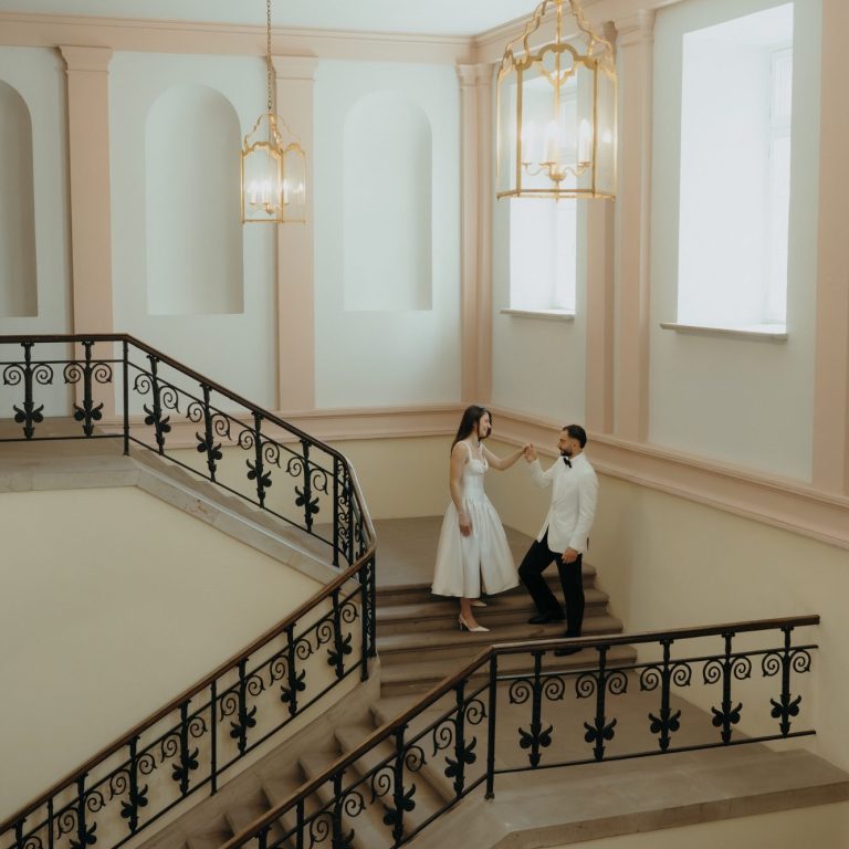 Ein Paar steht an einer Treppe in einem eleganten Innenraum. Helle Farben und Kunstlicht.