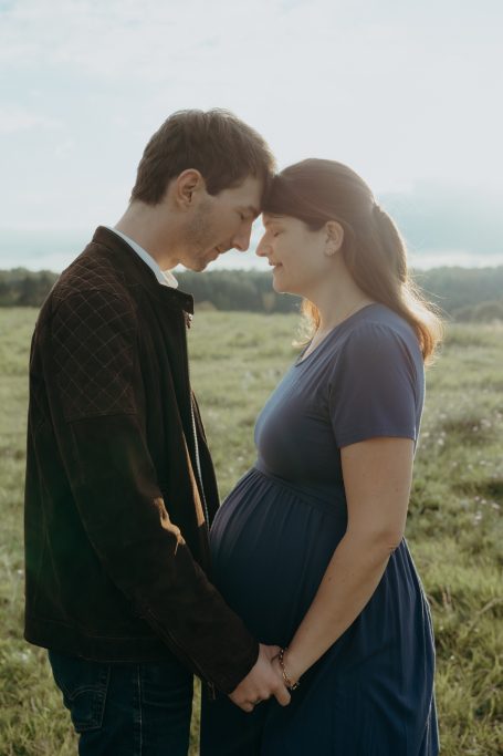 Ein Paar steht in einem Feld, die Hände auf dem Babybauch der Frau, sich lächelnd ansehend.
