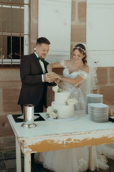 Ein Brautpaar schneidet gemeinsam eine Hochzeitstorte an einem Tisch an.