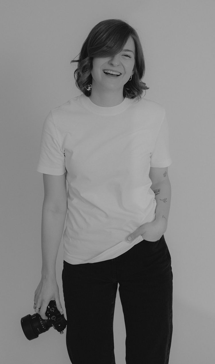 Lachende Person mit kurzen Haaren, die entspannt auf einen Stuhl gestützt ist.