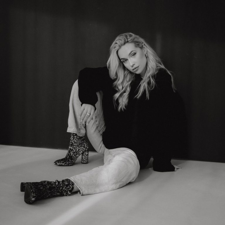 Eine blonde Frau sitzt lässig auf dem Boden in einem modischen Outfit.