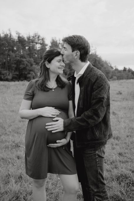 Ein Paar steht in einem Feld, sie umarmen sich und zeigt ihren Babybauch.