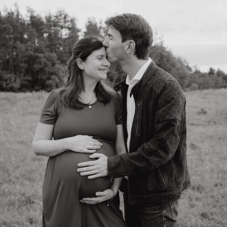 Schwangere Frau in einem Kleid steht mit einem Mann in einer Feldlandschaft.
