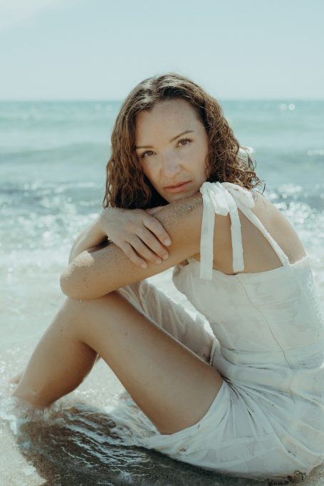 Frau mit lockigem Haar sitzt am Strand und schaut nachdenklich in die Kamera.