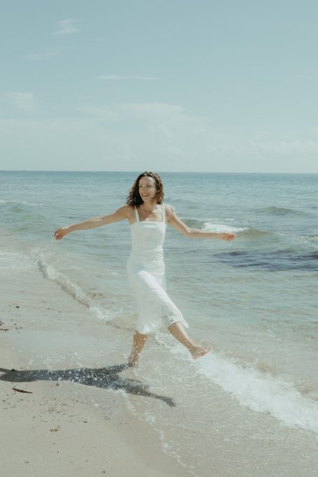 Frau in einem weißen Kleid, die fröhlich am Strand im Wasser spielt.